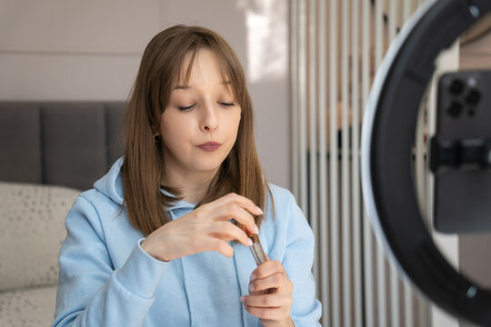 Young woman filming a beauty tutorial on her bed, applying lipstick under a ring light while holding a smartphone for social media makeup content and lifestyle vlogging
