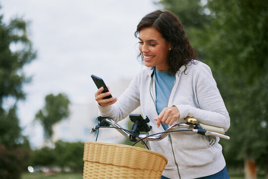 Woman smiling and interacting with her smartphone while standing next to her bicycle in a park, representing connection, leisure, and modern outdoor activity