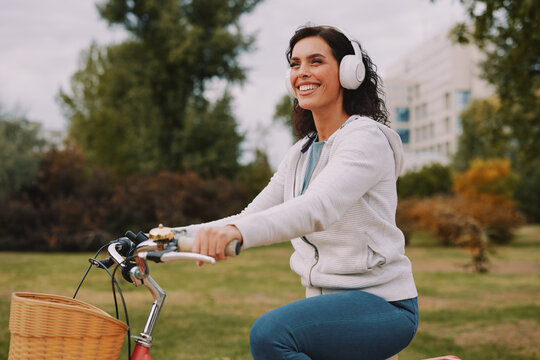 Happy woman with wireless headphones enjoying music while riding a bicycle in a city park, embracing leisure, wellness, and an active lifestyle outdoors