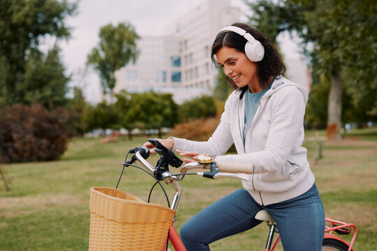 Woman riding a bicycle in a city park, wearing headphones and looking at a smartphone on the handlebar, enjoying music and navigating with technology for a healthy lifestyle