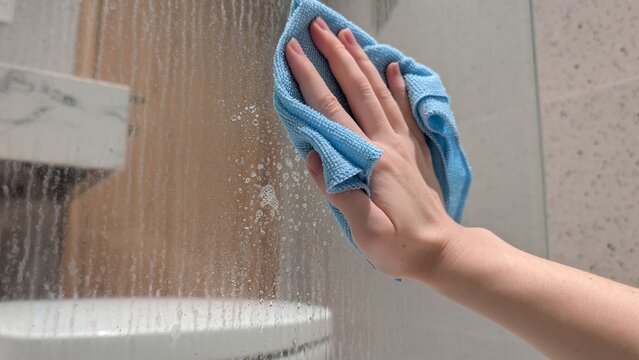 Woman's hand using a microfiber cloth and cleaning spray to wipe a dirty glass shower partition covered in hard water stains, limescale, and dried soap scum. Household chores and limescale removal.