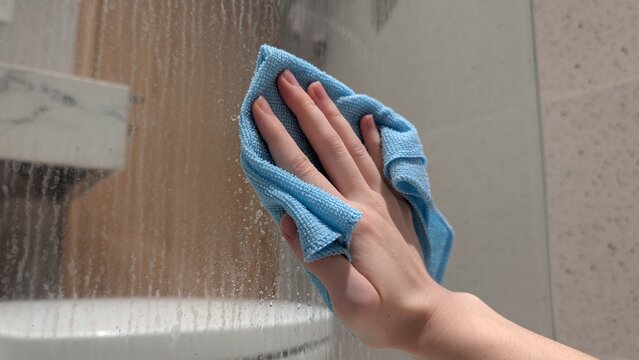 Woman's hand using a microfiber cloth and cleaning spray to wipe a dirty glass shower partition covered in hard water stains, limescale, and dried soap scum. Household chores and limescale removal.