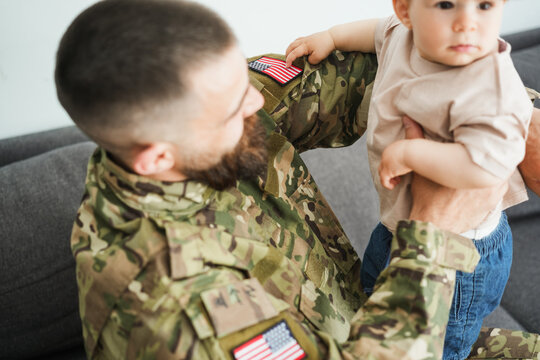 Male american soldier in military uniform hugs his little son at home while baby boy touching american US flag - Emotional military happy homecoming concept - Main focus on left flag - UGC content