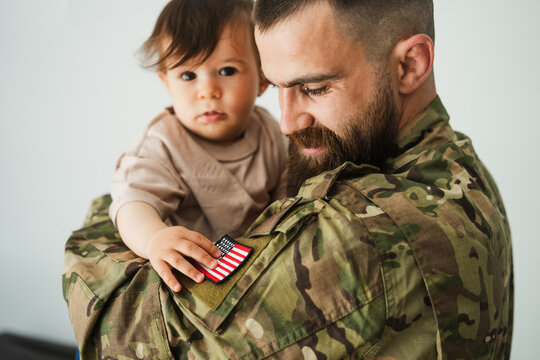 Male american soldier in military uniform hugs his little son at home while baby boy touching american flag - Emotional military happy homecoming concept - Main focus on man face - UGC content