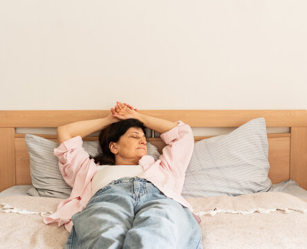Mature woman is lying on top of bedspread, her eyes closed and her arms stretched back behind her head. Mindful, unhurried lifestyle, slow living philosophy, conscious rest, relaxation, and self-care