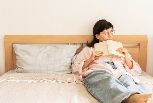 Mature woman in casual clothes and glasses is relaxing and reading book in bed at home. Slow living, mindful leisure, time for oneself, and calm lifestyle