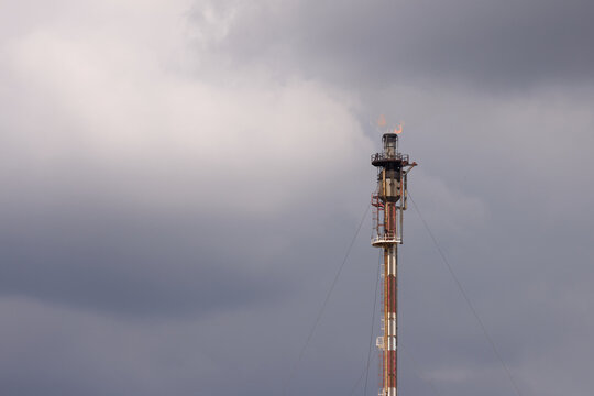 Industrial gas flare stack chimney burning hydrocarbon gases with bright orange flame at oil refinery against dark overcast clouds.