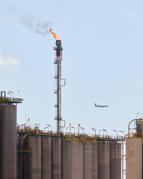 Industrial gas flare stack chimney burning fuel with orange fire and smoke over storage silos at oil refinery plant with airplane flying in sky.