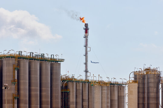 Industrial gas flare stack chimney burning fuel with orange fire and smoke over storage tanks at oil refinery plant with airplane flying in sky.
