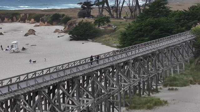 Aerial view of the historic wooden Pudding Creek Trestle bridge with people walking across above the sandy beach and ocean waves in Fort Bragg, California, United States.
