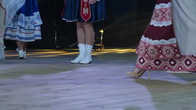 Low angle view captures female feet wearing white boots and heels dancing in vibrant ethnic skirts and embroidered folk dresses across wooden stage