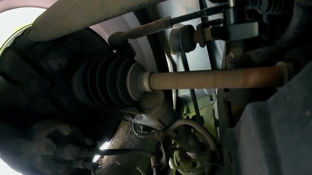 Front axle rotating beneath moving vehicle while suspension components respond to road movement. CV boot and drive shaft visible beside steering tie rod. Close mechanical view of wheel hub assembly