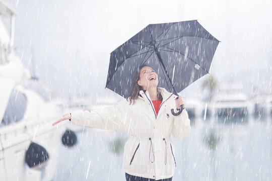 Joyful Woman in White Coat with Black Umbrella Enjoying Rain on a Waterfront