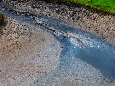 Aerial view of a dried-up riverbed with dark, wet mud and sediment, surrounded by patches of green grass in Latvia.