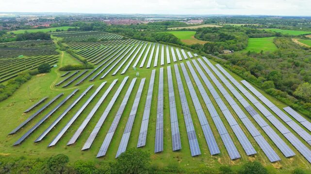 Photovoltaic solar farm across open green fields with long rows of panels. Rural renewable energy site near woodland and farmland. Large scale clean electricity infrastructure under cloudy sky