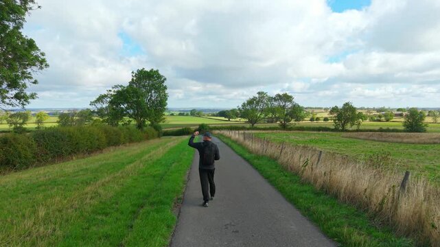 Backpacker walking along rural road between green fields and hedgerows. Active lifestyle and countryside hiking in open farmland. Male tourist exploring rural landscape on foot with backpack under