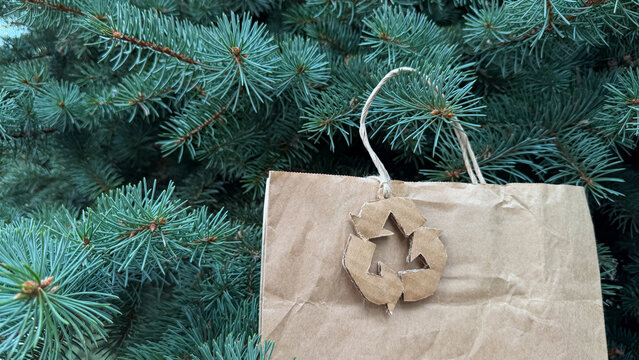 Craft paper gift bag with cardboard recycling logo on fir tree, eco christmas shopping, holiday delivery.