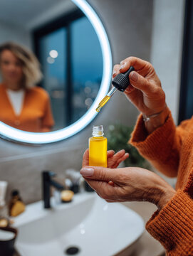 A woman is applying a serum to her hand in the bathroom, with a mirror reflecting herself and another person. She has manicured nails painted pinkish color.