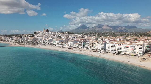 The camera captures the Albir and Altea region along the Costa Blanca coastline. Sun shines down on the beach and clear water. People walk and relax by the shore.