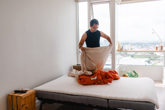 Asian man making bed in modern apartment bedroom with city view, performing daily chores.