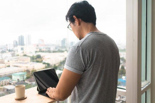 Asian man working remotely on a digital tablet with a keyboard by a city window.