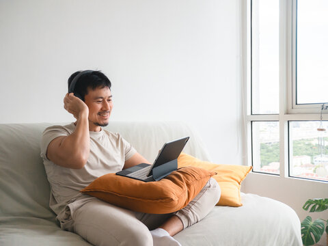 Smiling Asian man wearing headphones, relaxing on sofa while using digital tablet at home