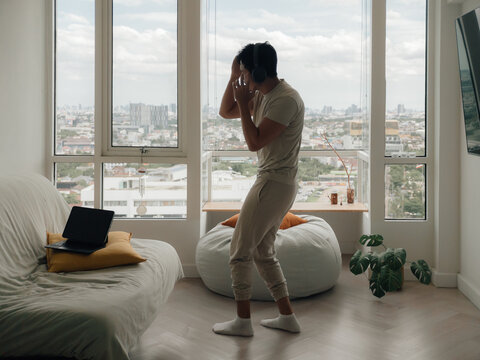 Young man wearing headphones dancing energetically in his modern apartment with a city view.