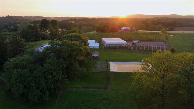 Aerial view of Flintrock farm at sunset, featuring greenhouses, a small pond, lush green fields, and rolling hills in Elizabeth Township, Pennsylvania, United States.