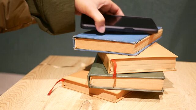 A person grabs mobile phone from top of stack of books, showing educational content and modern technology on wooden table.