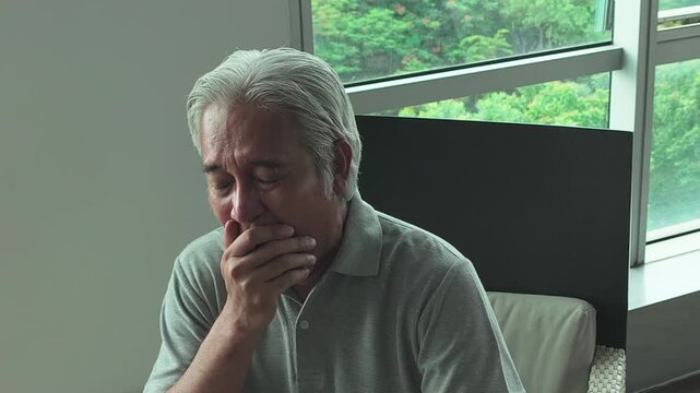 middle-aged southeast asian man relaxing in armchair, yawning and feeling sleepy indoors at home