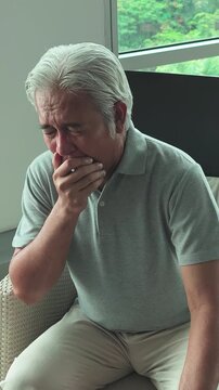 middle-aged southeast asian man relaxing in armchair, yawning and feeling sleepy indoors at home