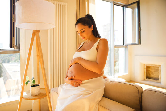 Pregnant woman in white dress gently cradles her belly while sitting on a beige sofa in a bright living room with large windows and a floor lamp