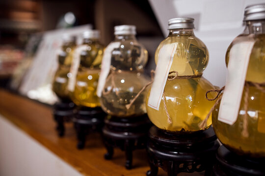 Decorative glass bottles filled with yellow liquid displayed on wooden shelf with black stands in a modern interior setting, labels attached for identification