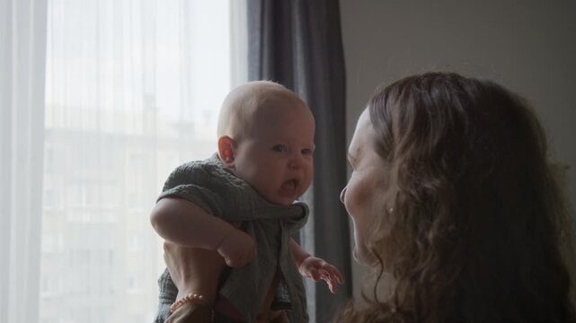 Caucasian young adult mother lifting infant baby, talking and smiling near window. Footage suitable for maternal healthcare campaigns, family parenting commercials, baby product advertising