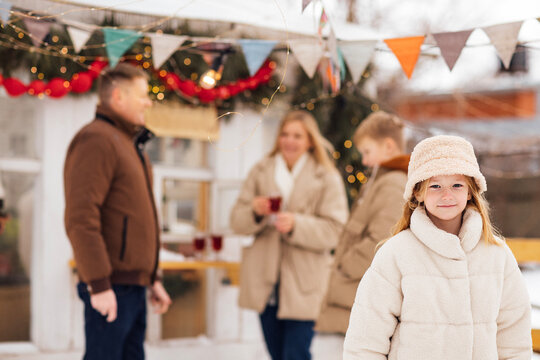 Young girl in white winter coat smiles at the camera while family members enjoy drinks in a festive outdoor setting with decorations and snow