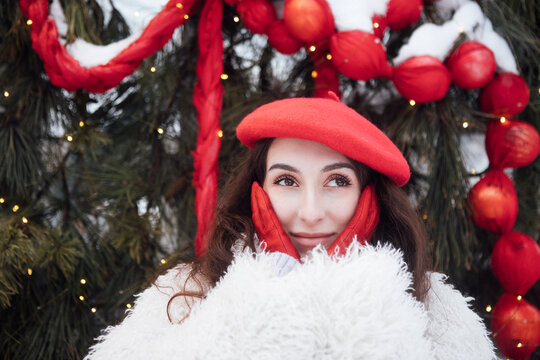 Young woman wearing a red beret and white fluffy coat poses with hands on her cheeks, surrounded by festive decorations and evergreen branches in a winter setting