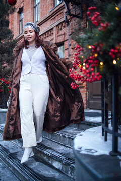 Fashionable woman in a brown fur coat and white outfit walks down snowy steps decorated with holiday greenery and red berries in a winter urban setting