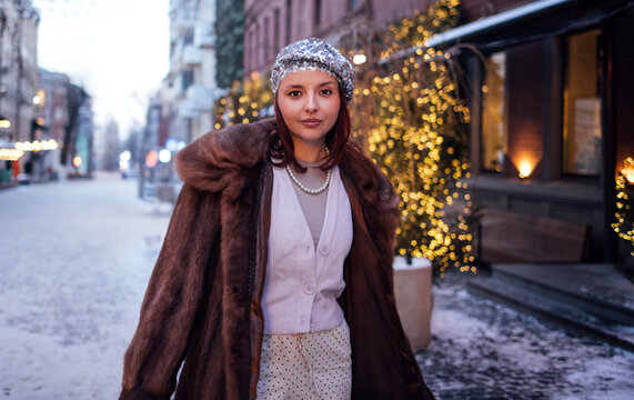 Young woman wearing a stylish fur coat and silver headband walks along a snow-covered street adorned with festive lights and decorations in a city setting