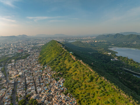 Aerial view of the historic city of Udaipur, featuring a prominent green hill ridge separating dense urban rooftops from the serene lake under a clear blue sky. Udaipur, Rajasthan, India.