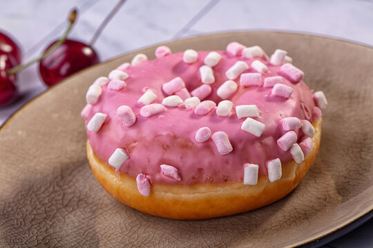 Pink glazed donut topped with mini marshmallows served on textured ceramic plate with fresh cherries in background