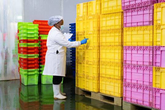 Quality control officer with clipboard checking stock in industrial warehouse, worker in protective wear inspecting plastic crates in manufacturing plant