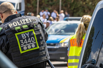 German police officers in high-visibility yellow vests stand outside next to a patrol car during an operation. In the background, more police vehicles are visible on the street during daytime. © Thomas