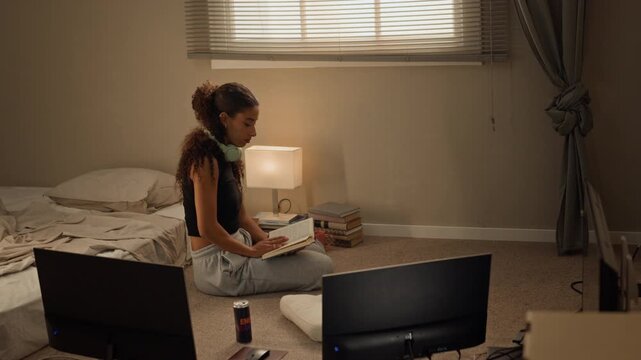 Multiethnic young woman sitting cross-legged on floor reading notebook with headphones around neck. Footage ideal for e-learning apps, study campaigns and remote education commercials