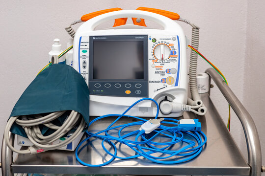 Illustrative editorial image of a Nihon Kohden Cardiolife automated external defibrillator device resting on a stainless steel hospital cart with medical cables.