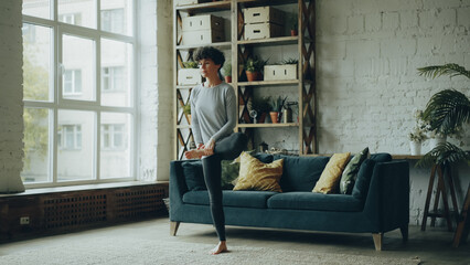Slim yogini woman is doing yoga alone in modern loft style apartment balancing on one leg with...