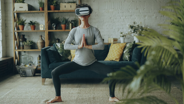 Cute flexible girl is doing stretching exercises at home wearing artificial reality glasses enjoying amazing experience using modern device. Sports and technology concept.