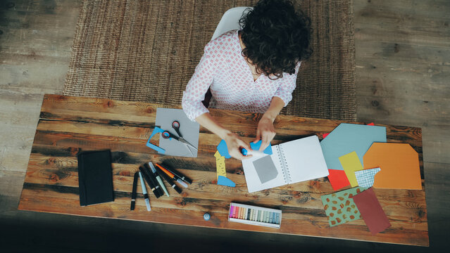 Top view of busy female designer sticking paper figure in notebook using glue-stick working at home alone at table. Creativity, design and modern art concept.