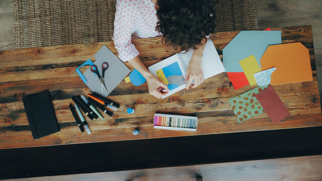 Zoom-in of female employee creative designer drawing on splash-paper collage in notebook with crayons sitting at table in modern office. Job, people and creativity concept.