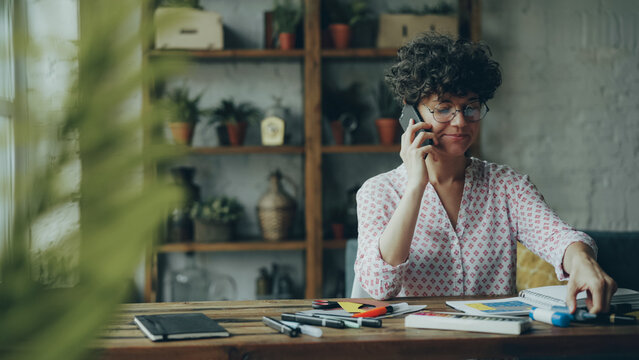Attractive girl designer is talking on smartphone sitting at table and touching stationery working at home. Modern communication, youth and workplace concept.