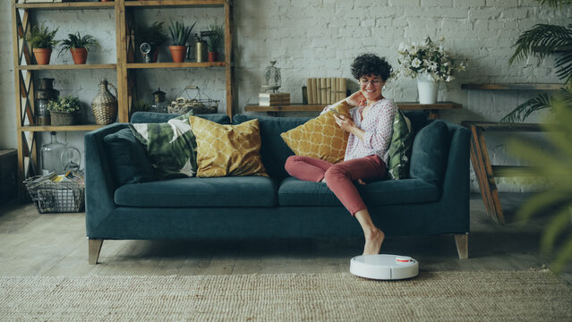 Joyful young woman is playing with robotic vacuum cleaner then using smartphone sitting on couch at home enjoying modern technology. People, household and innovations concept.